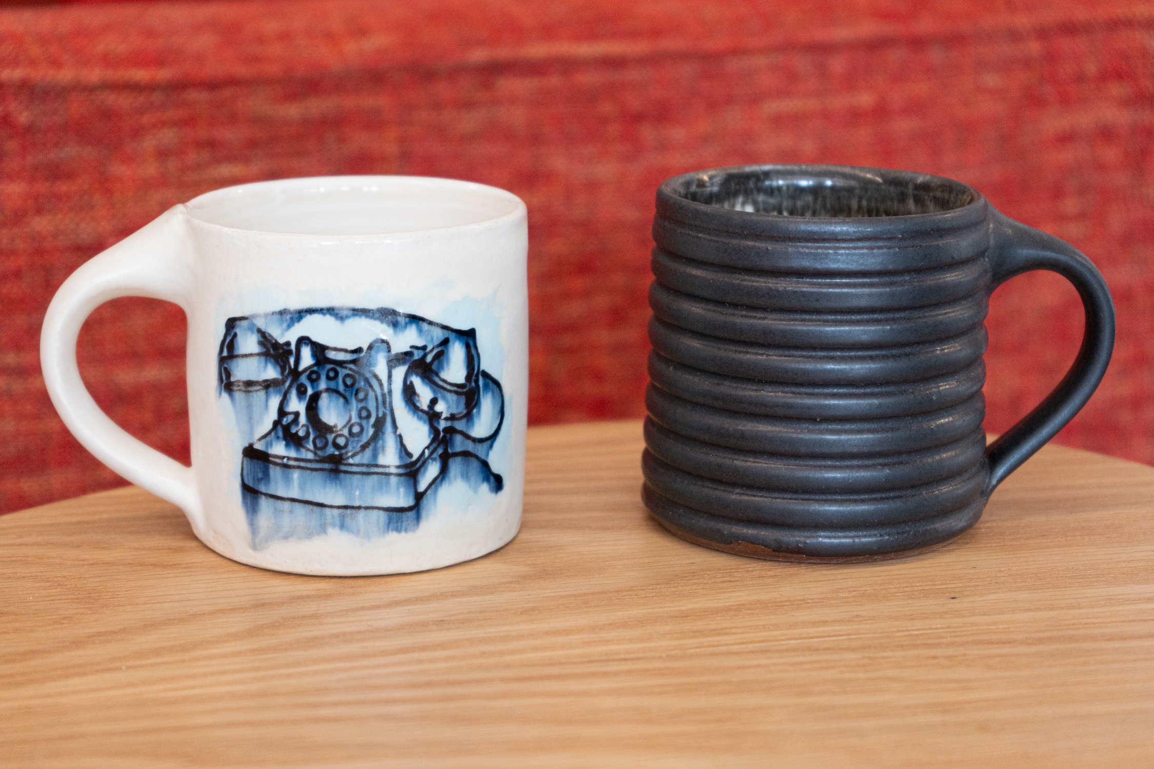 Two wheel thrown mugs on a light wood table with a red background