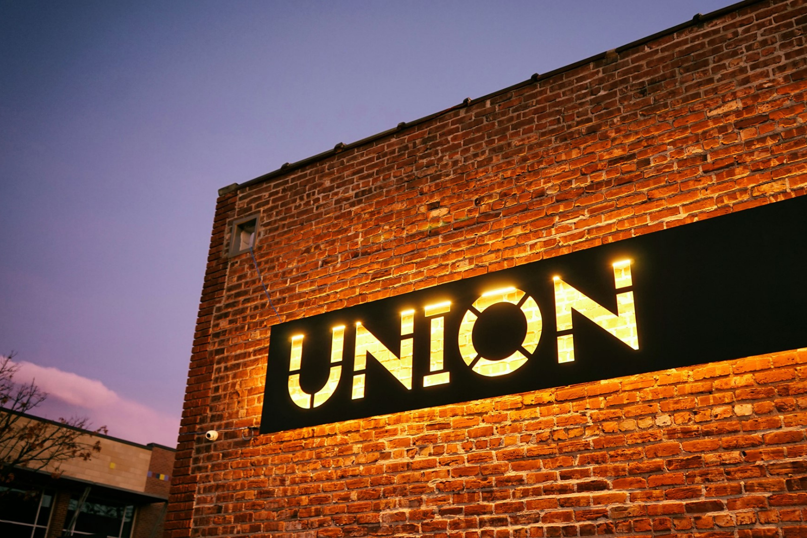 Union building at dusk A black steel Union sign hangs on a red brick wall and is illuminated with white light from behind